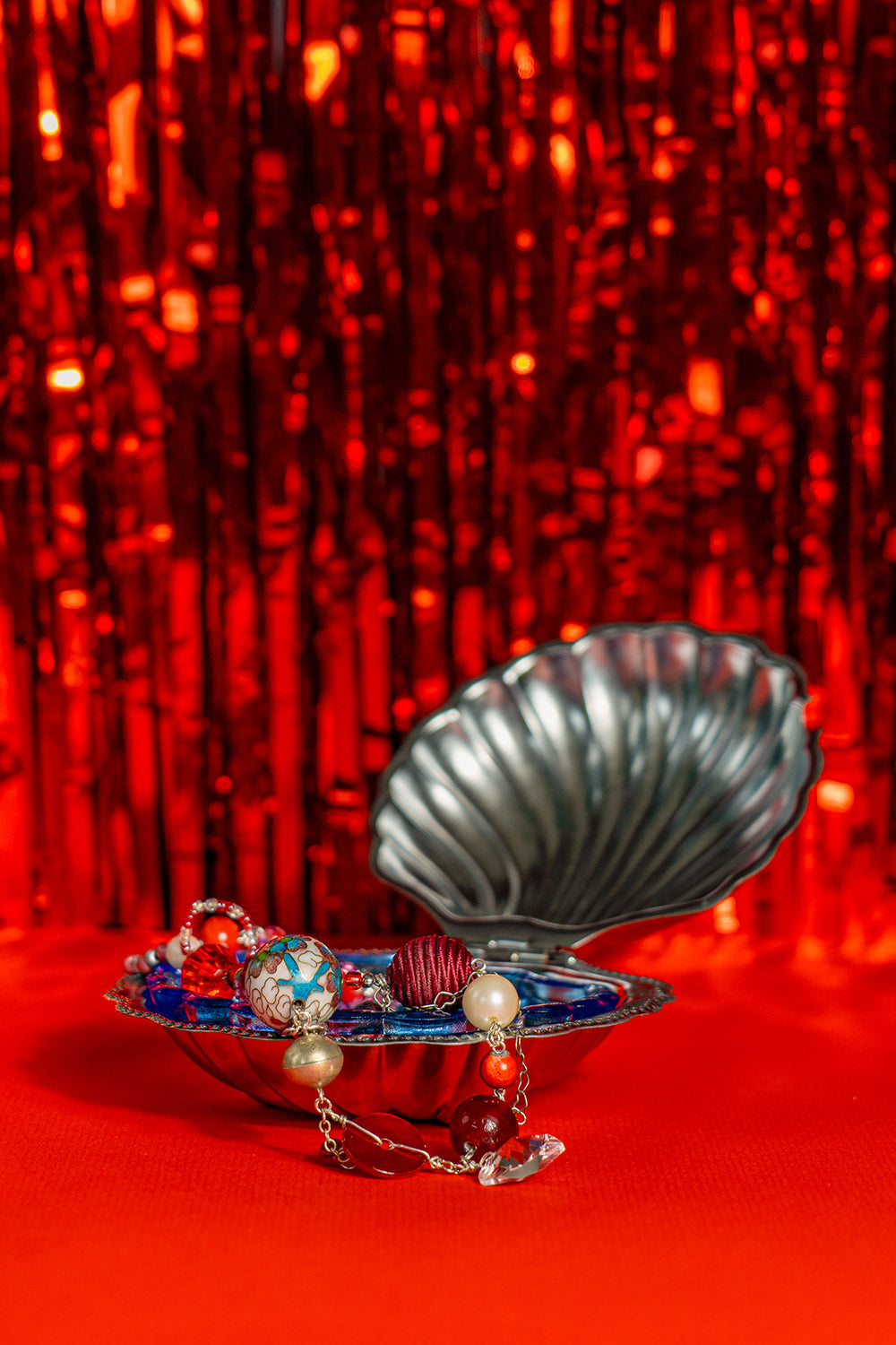A vintage seashell butter dish used as a trinket dish, holding a colorful handmade bead necklace. They sit on a bright red tinfoil background.