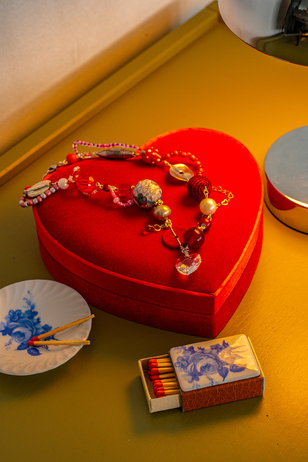 A colorful handmade bead necklace sitting on a heart shaped box, next to a vintage set of porcelain matchbox and small plate.