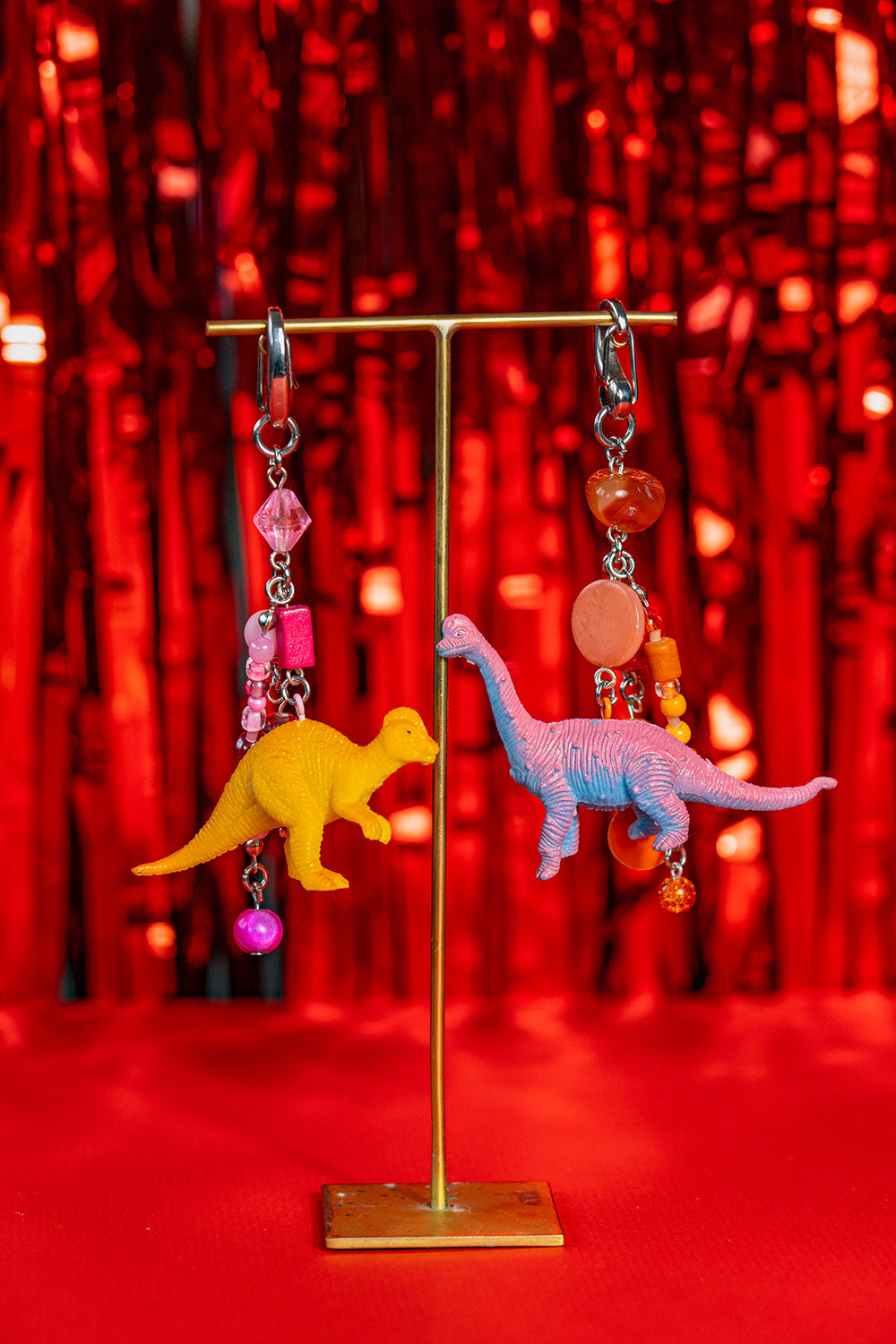 A pair of handmade matching keychains on a jewelry stand, made with miniature toy figurine dinosaurs. One has pink beads and an orange dino, the other orange beads and a pink dino.  