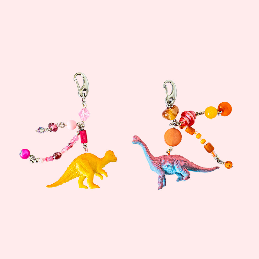 A pair of handmade matching keychains, made with miniature toy figurine dinosaurs. One has pink beads and an orange dino, the other orange beads and a pink dino.  