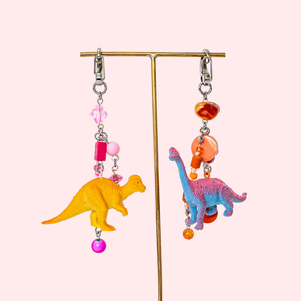 A pair of handmade matching keychains on a jewelry stand, made with miniature toy figurine dinosaurs. One has pink beads and an orange dino, the other orange beads and a pink dino.  
