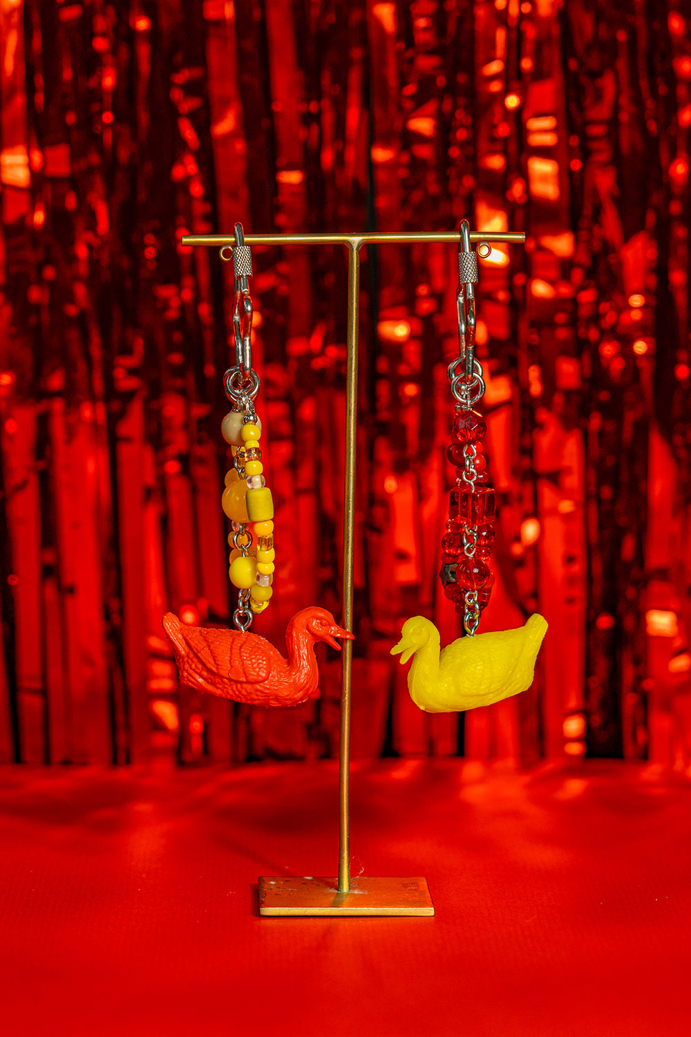 A pair of handmade matching keychains on a jewelry stand, made with miniature toy figurine geese. One has yellow beads and a red goose, the other red beads and a yellow goose.  