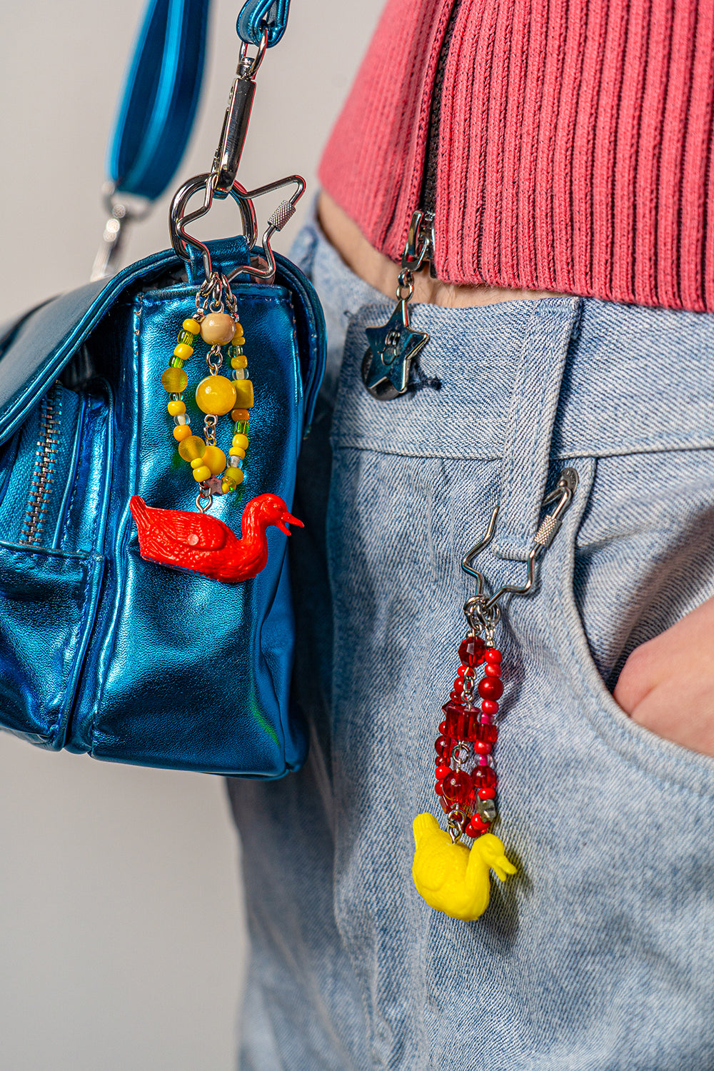 A pair of handmade matching keychains, made with miniature toy figurine geese. One has yellow beads and a red goose and is worn on a bag, the other red beads and a yellow goose and is worn on a belt loop of some denim pants.