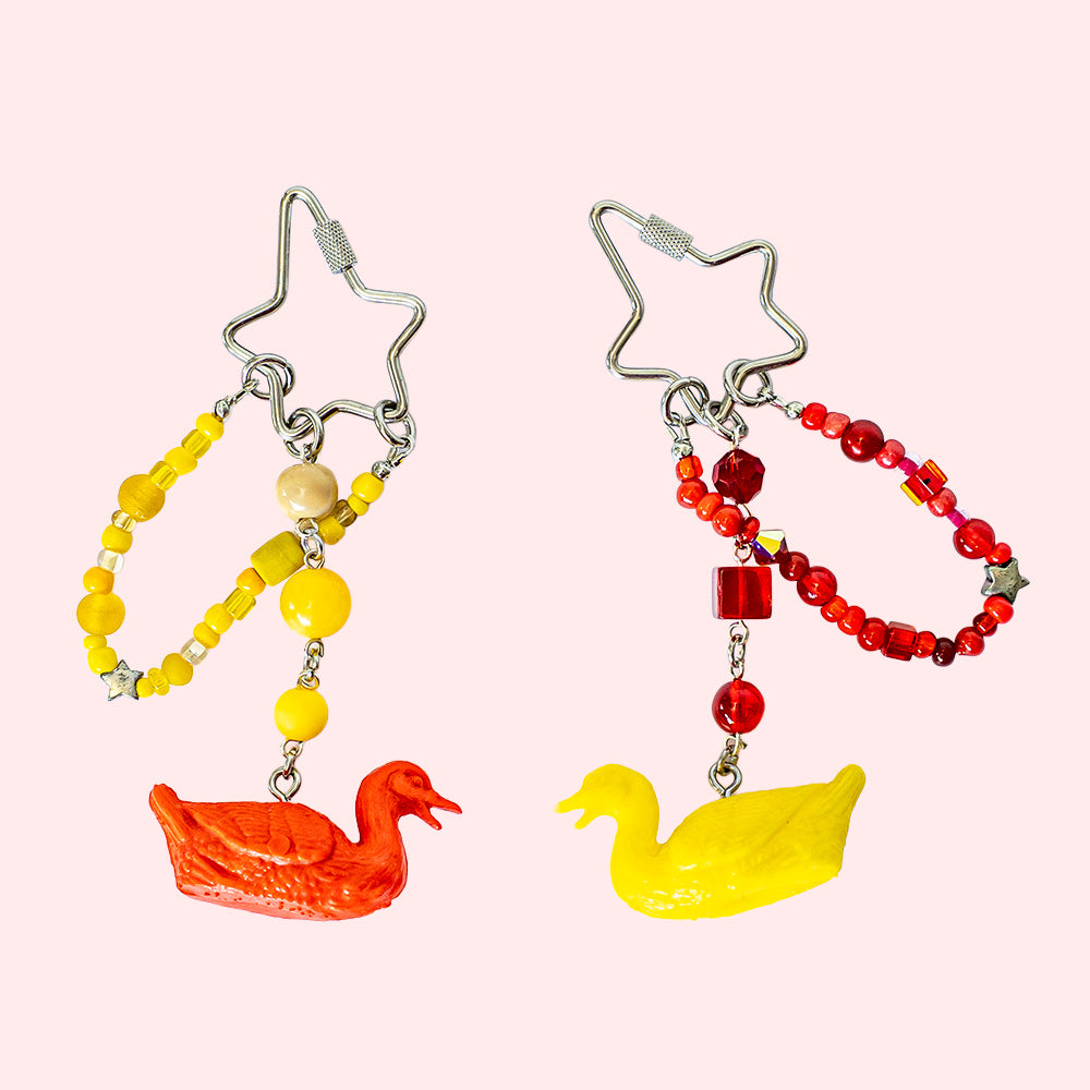 A pair of handmade matching keychains made with miniature toy figurine geese. One has yellow beads and a red goose, the other red beads and a yellow goose.  