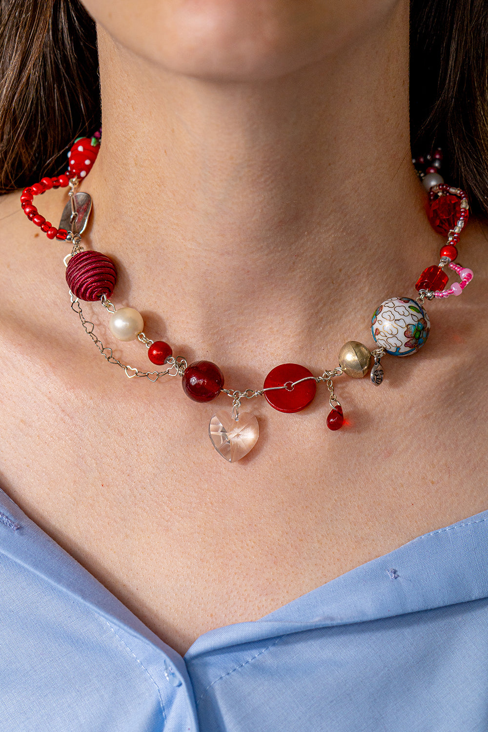 Handmade, colorful (mostly red) necklace in silver and red made with large beads of various materials. In the middle is a glass heart pendant. It's worn by a person in a closeup.