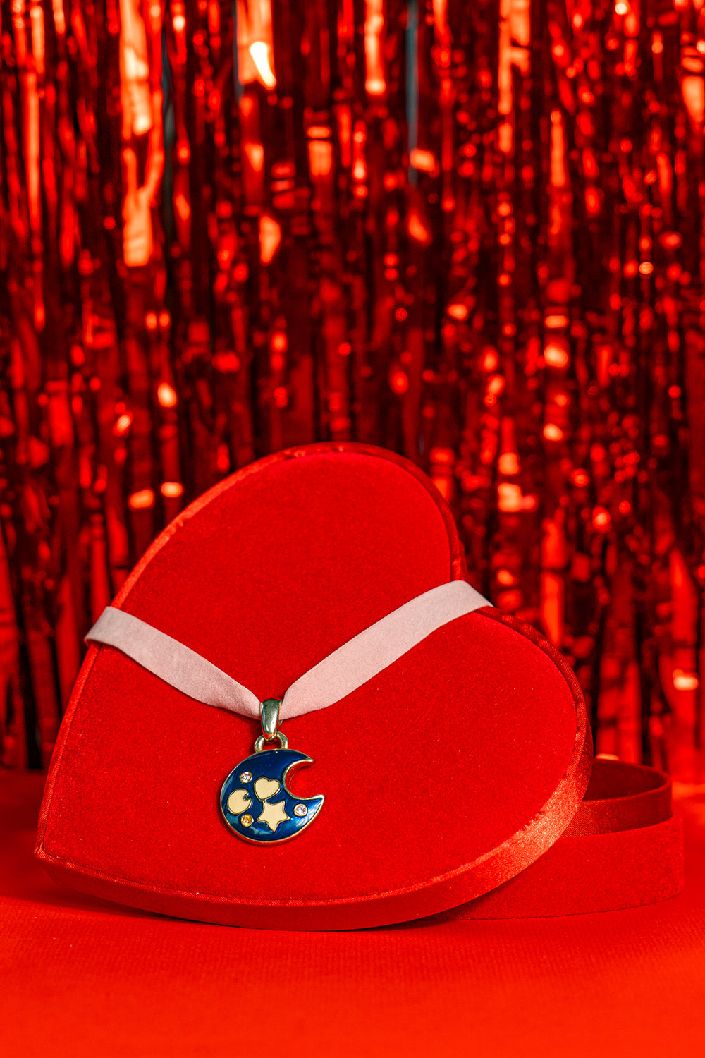 A choker in pink ribbon with a large pendant representing a dark blue moon crescent with small white symbols, sitting on a heart shaped box in front of a bright red tinsel background.