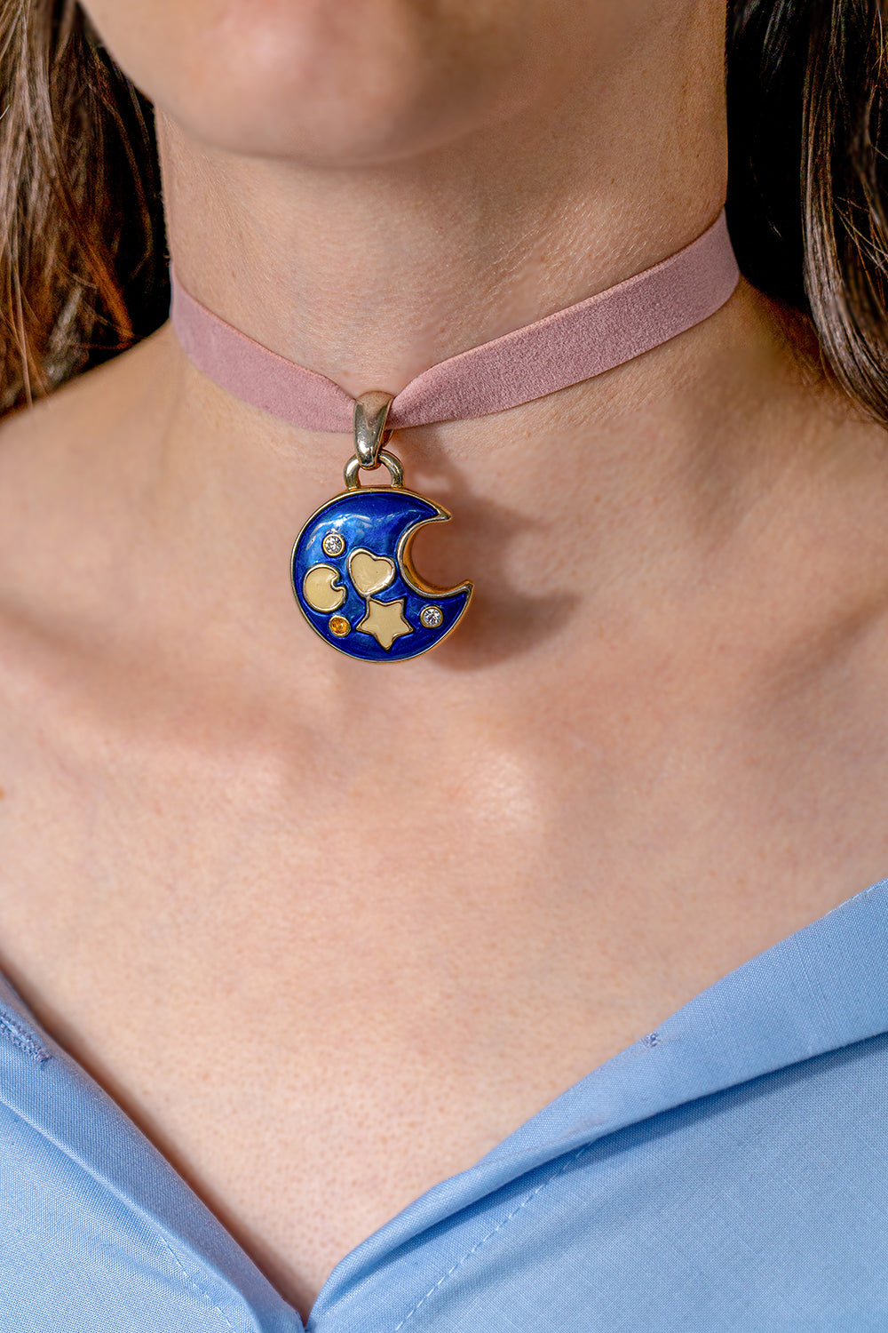 A choker in pink ribbon with a large pendant representing a dark blue moon crescent with small white symbols. It is worn tight around someone's neck.
