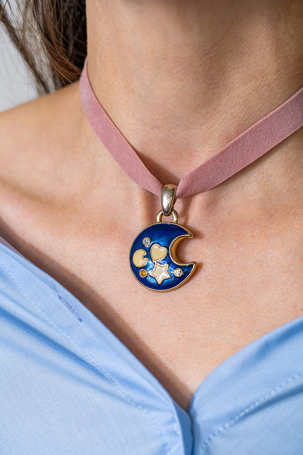 A choker in pink ribbon with a large pendant representing a dark blue moon crescent with small white symbols. It is worn slightly loose around someone's neck.