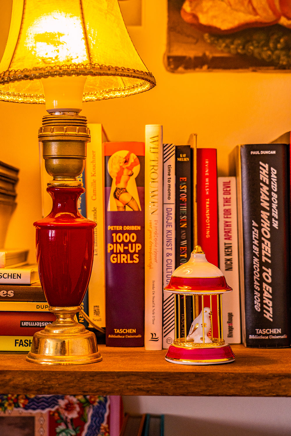 A vintage Limoges porcelain white, golden and red birdcage with a small white and golden bird sitting in it. It is on a bookshelf next to a lamp.