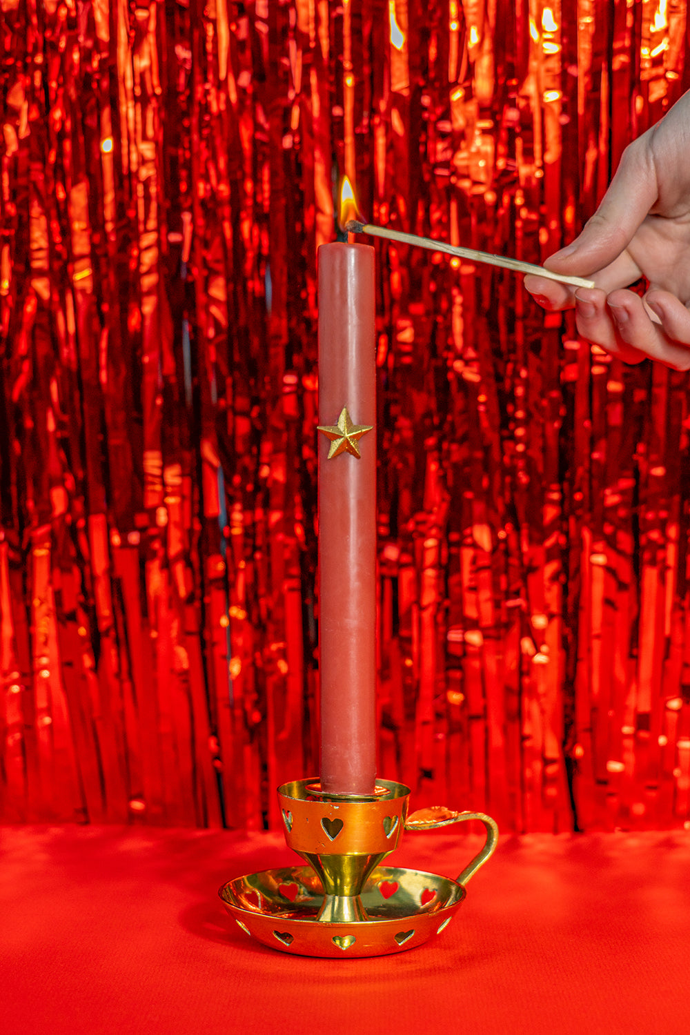 A golden vintage candlestick with heart cutouts around the base and the candle holding part. It also has a heart on top of the handle. it holds a red candle being lit, on a bright red background.