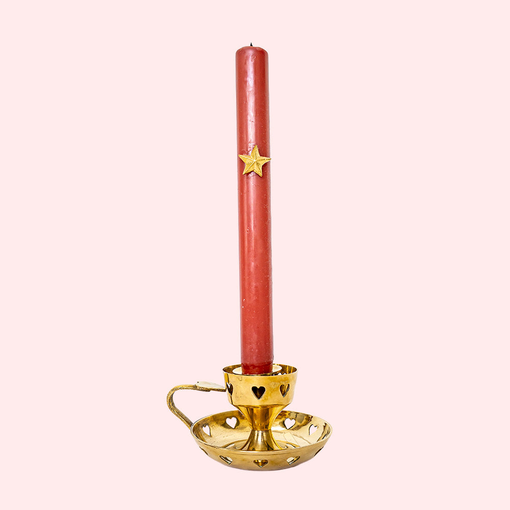 A golden vintage candlestick with heart cutouts around the base and the candle holding part. It also has a heart on top of the handle. It's holding a tall red candle.
