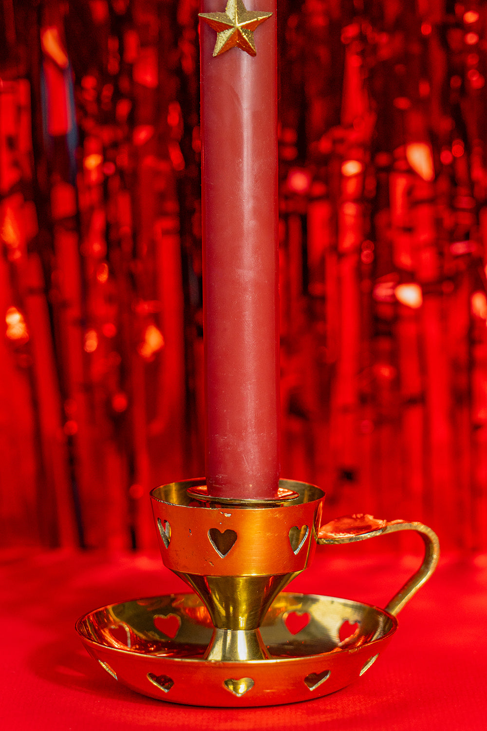 A golden vintage candlestick with heart cutouts around the base and the candle holding part. It also has a heart on top of the handle. it holds a red candle, on a bright red background.