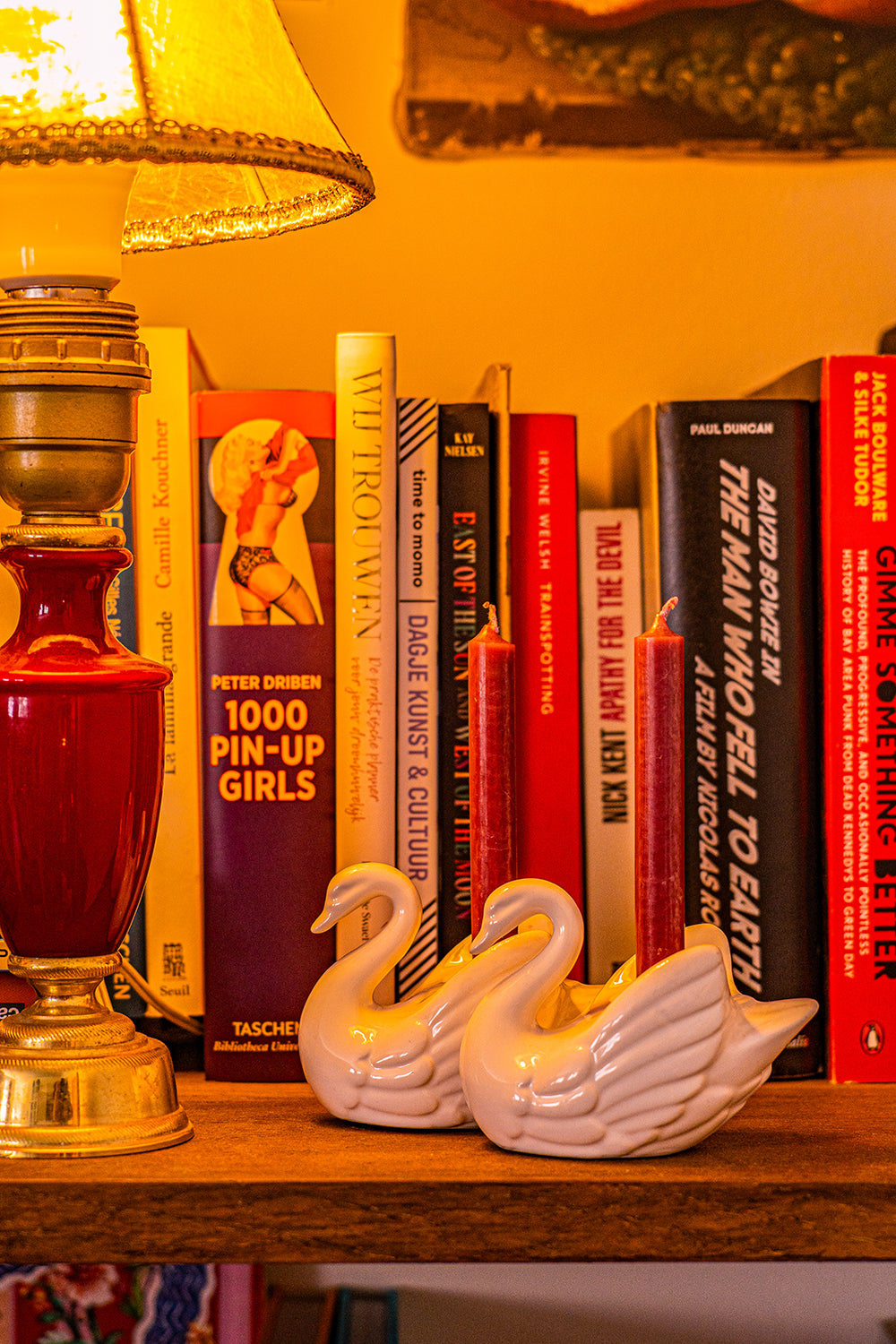 A vintage porcelain candlestick in the shape of a couple of swans, each holding a small red candle between their wings. It is on a bookshelf next to a lamp.