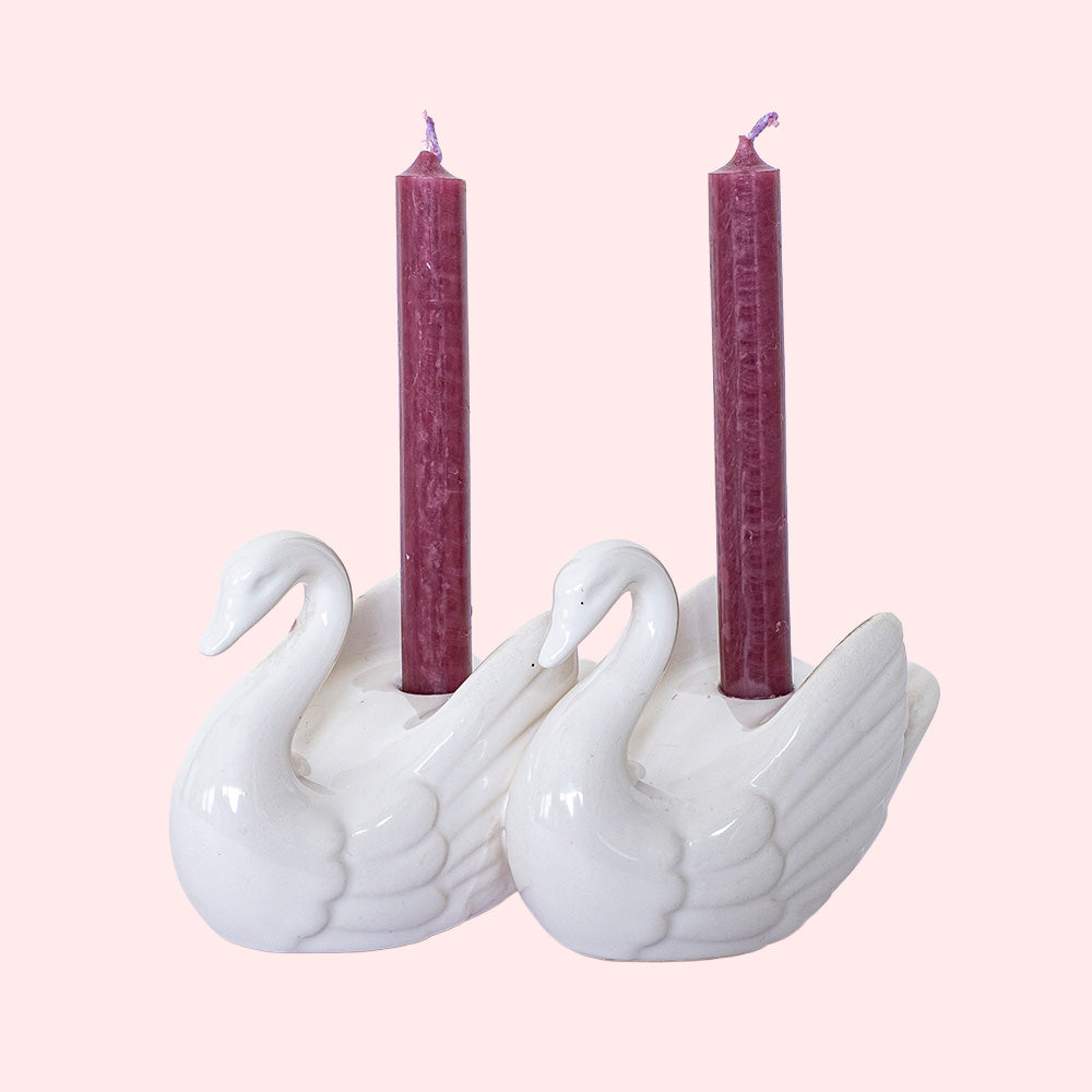 A vintage porcelain candlestick in the shape of a couple of swans, each holding a small red candle between their wings.