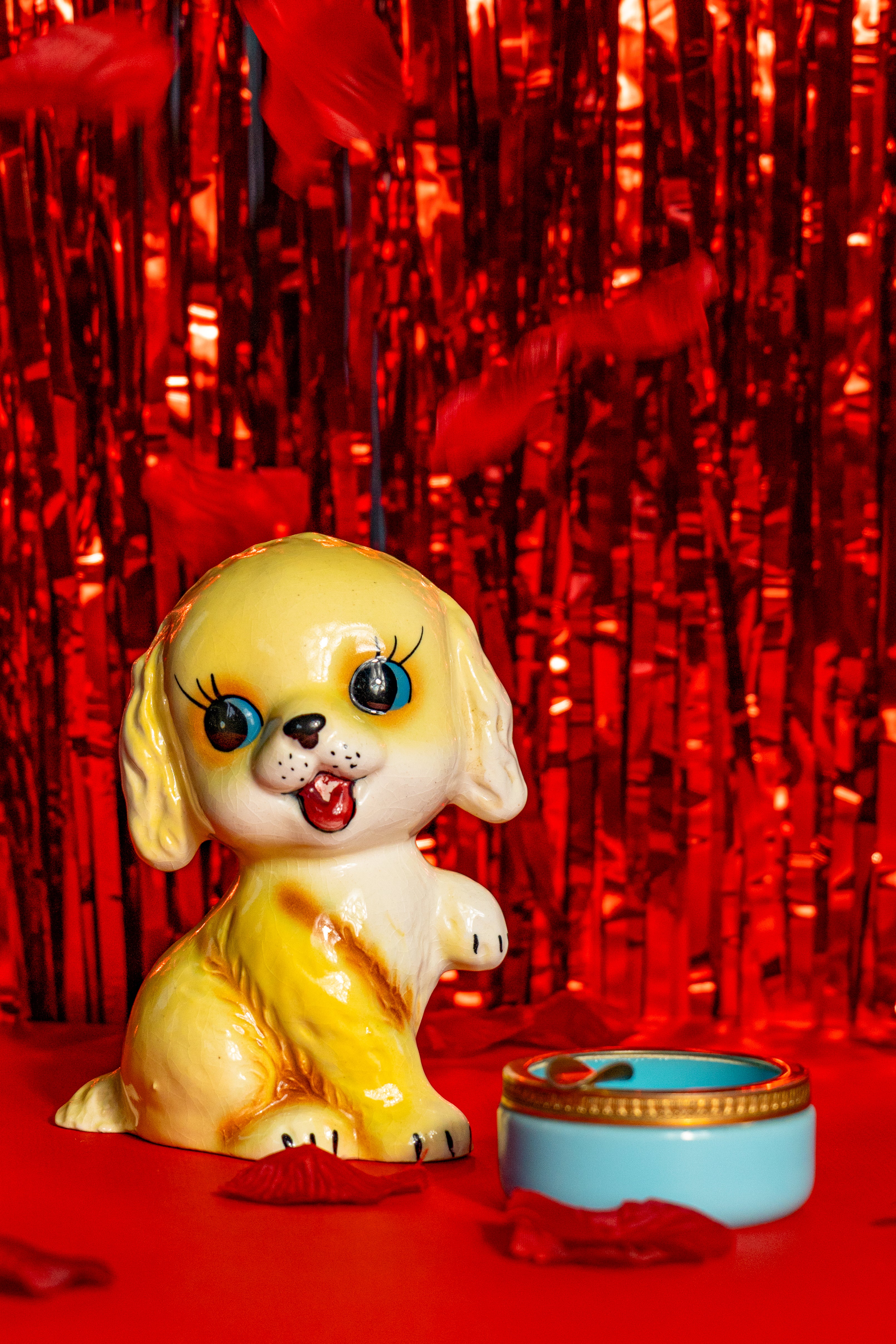 Second-hand Japan-style ceramic piggy bank from the 50's, representing a golden retriever with big eyes. Next to a vintage opaline Murano ashtray. Both are on a bright red tinfoil background and have rose petals falling on them.