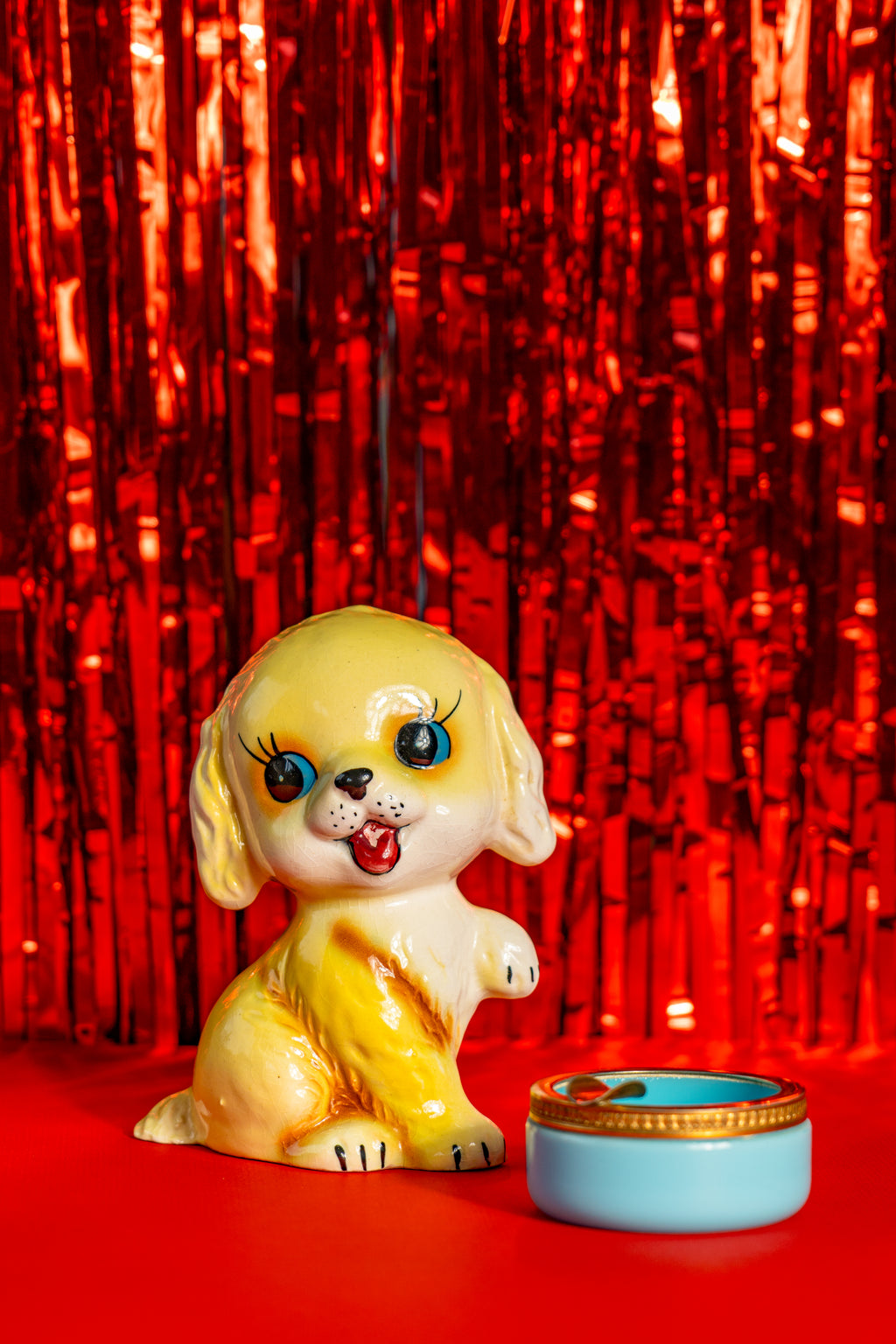Second-hand Japan-style ceramic piggy bank from the 50's, representing a golden retriever with big eyes. Next to a vintage opaline Murano ashtray. Both are on a bright red tinfoil background.