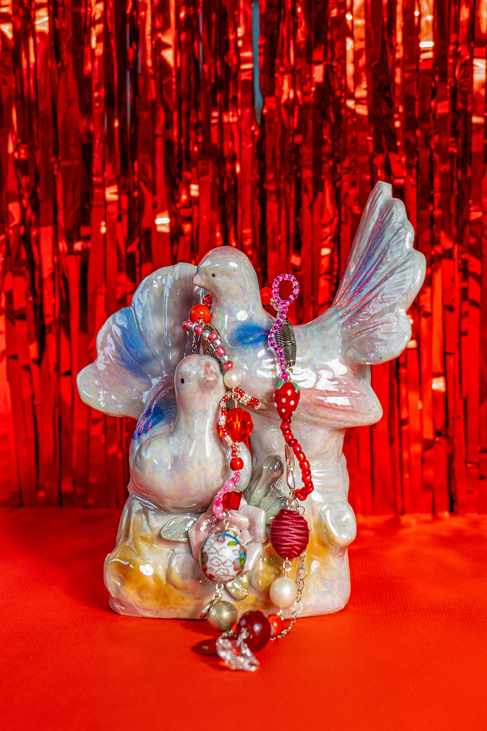 Second-hand, decorative ceramic figurine representing two doves with colorful wings and tails. It has an iridescent glaze. It sits on a bright red background and on it is laying a handmade necklace made from various beads.