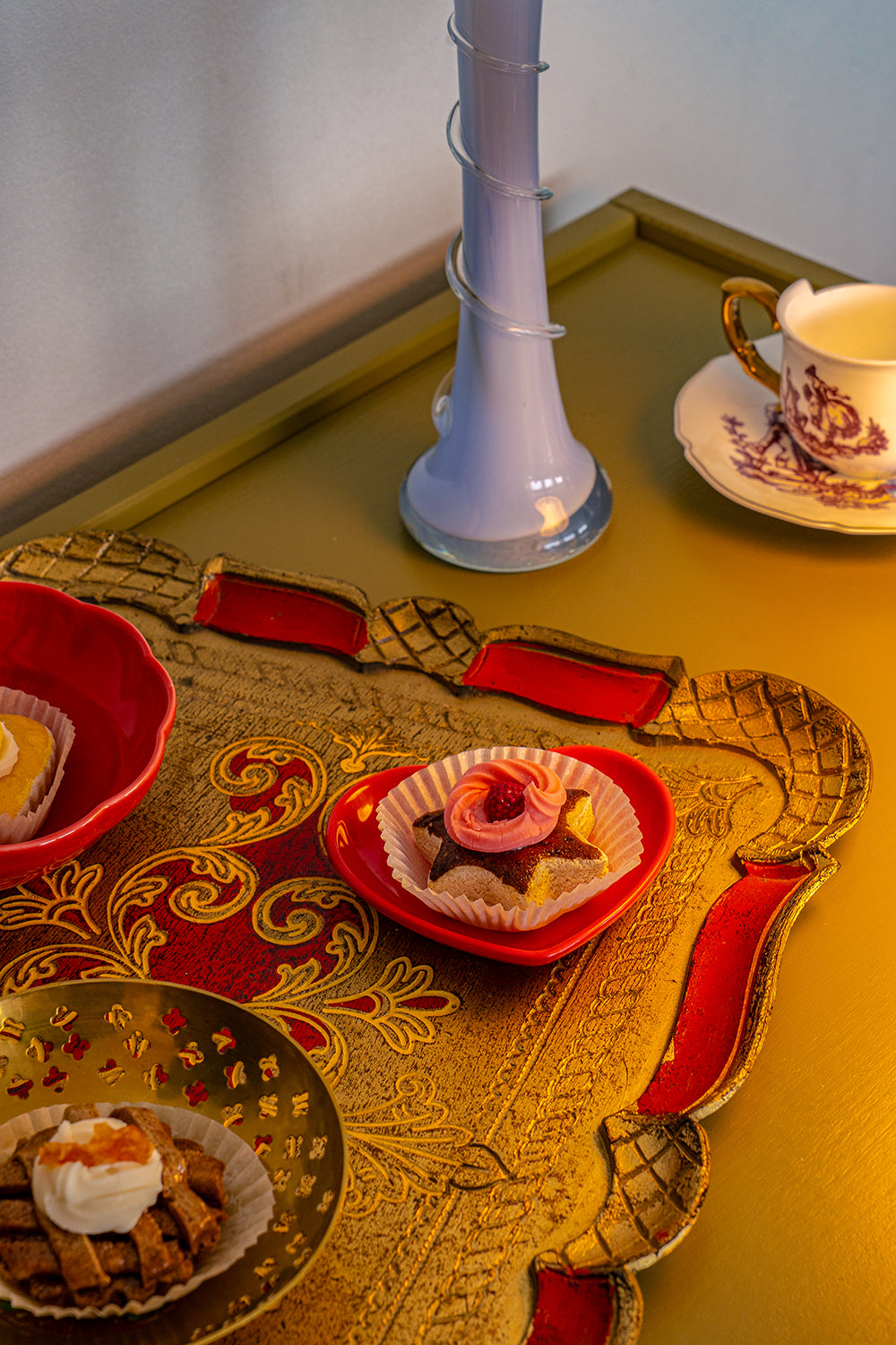 A decorative wooden tray painted red and golden with small cakes on it, in front of a blue opaline vase.