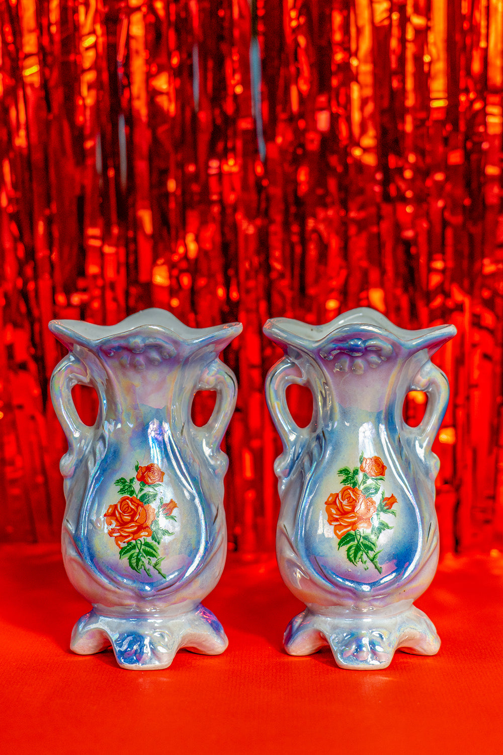 Two identical vintage small vases with a foot and handles on the side. They are two shades of blue with a print of roses on the front and an iridescent glaze. 