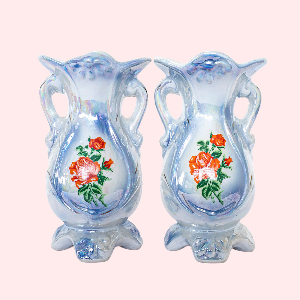 Two identical vintage small vases with a foot and handles on the side. They are two shades of blue with a print of roses on the front and an iridescent glaze. 