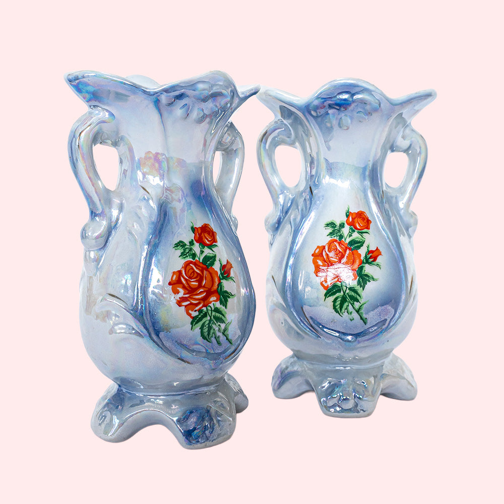 Two identical vintage small vases with a foot and handles on the side. They are two shades of blue with a print of roses on the front and an iridescent glaze. 
