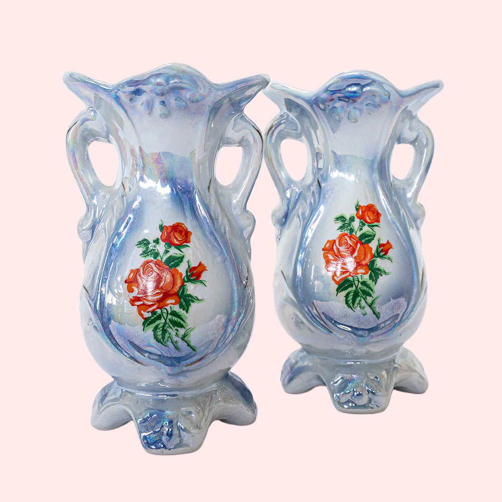 Two identical vintage small vases with a foot and handles on the side. They are two shades of blue with a print of roses on the front and an iridescent glaze. 