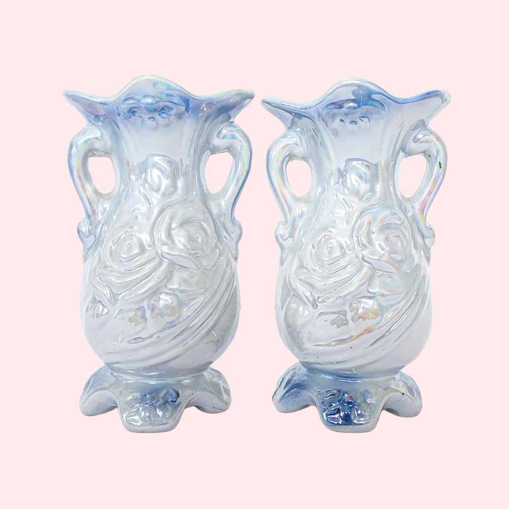 Two identical vintage small vases with a foot and handles on the side. They are two shades of blue with a print of roses on the front and an iridescent glaze. Seen from behind.