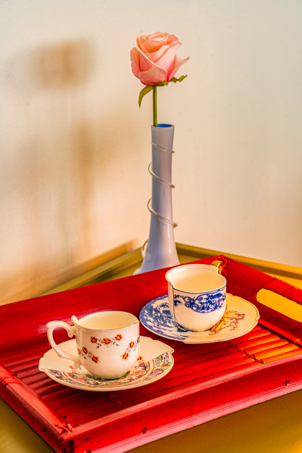 A red bamboo tray with two teacups on it and a small blue opaline vase holding a pink rose behind it.