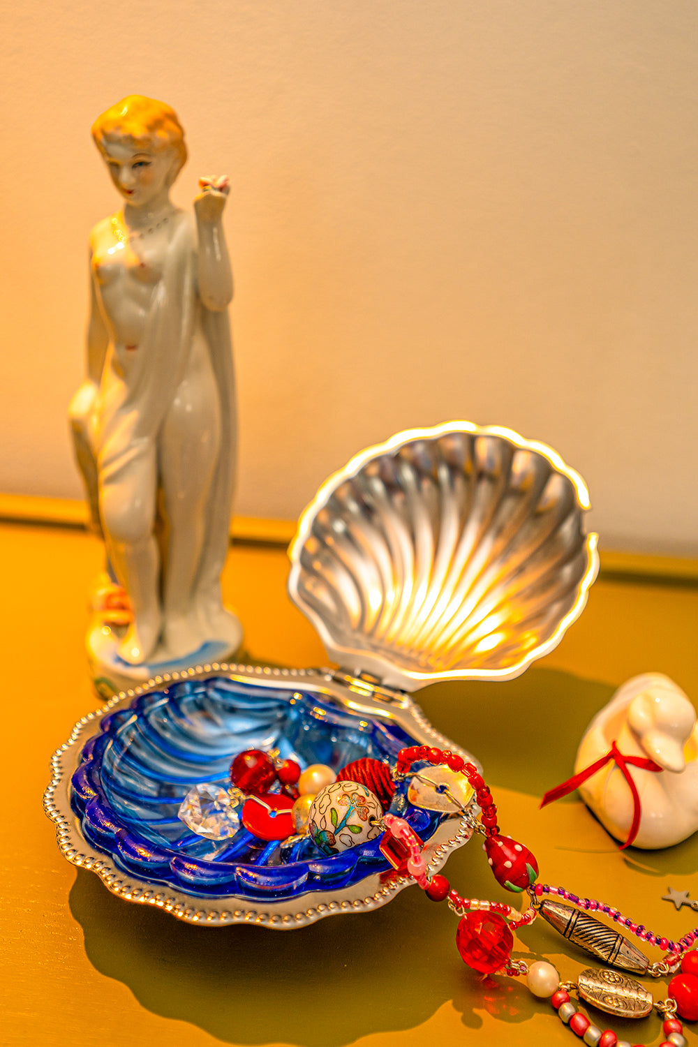 A vintage seashell butter dish used as a trinket dish, holding a colorful handmade bead necklace. They are on a table between a small statue of a naked lady and a small duck figurine.