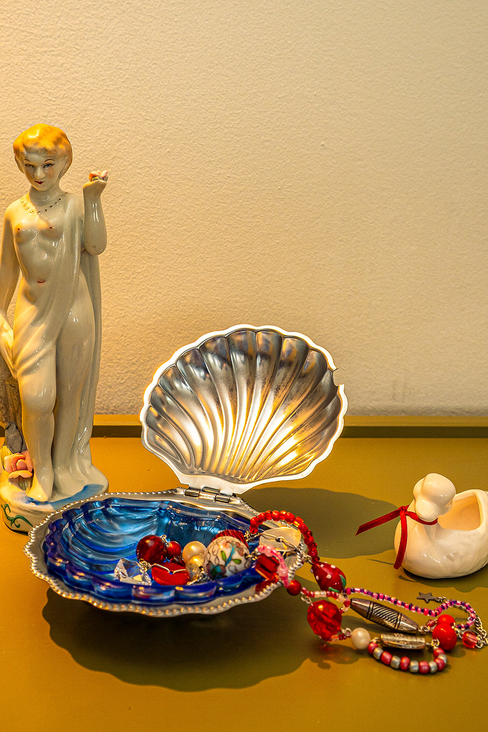A vintage seashell butter dish used as a trinket dish, holding a colorful handmade bead necklace. They are on a table between a small statue of a naked lady and a small duck figurine.