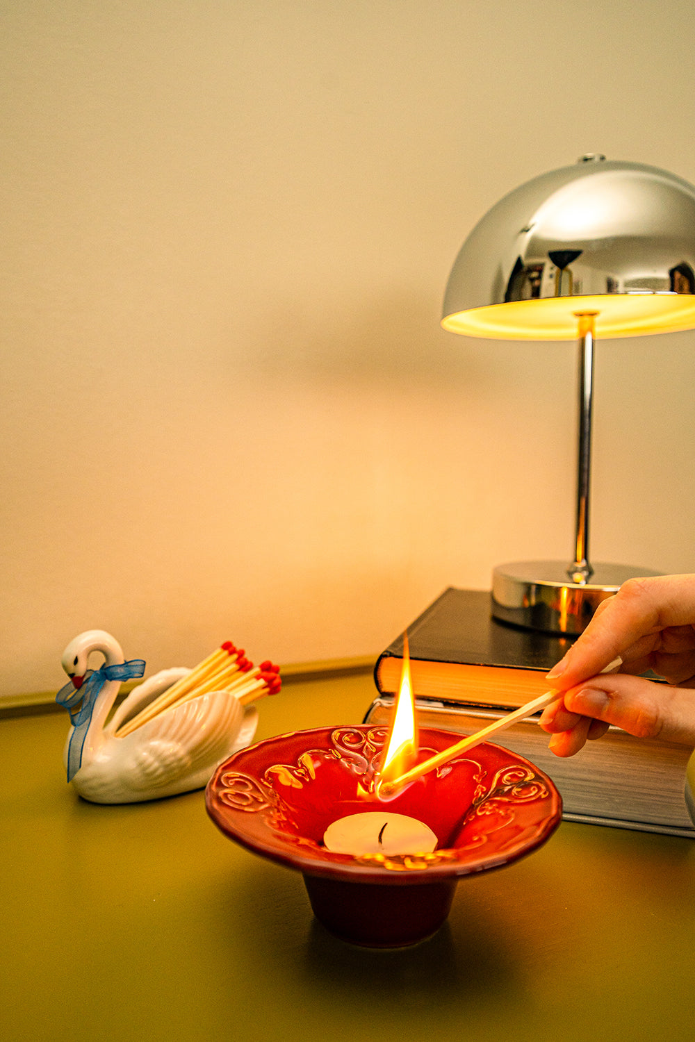 Second hand, ceramic figurine swan with matches on a table next to a red candleholder with a tealight being lit by a hand.