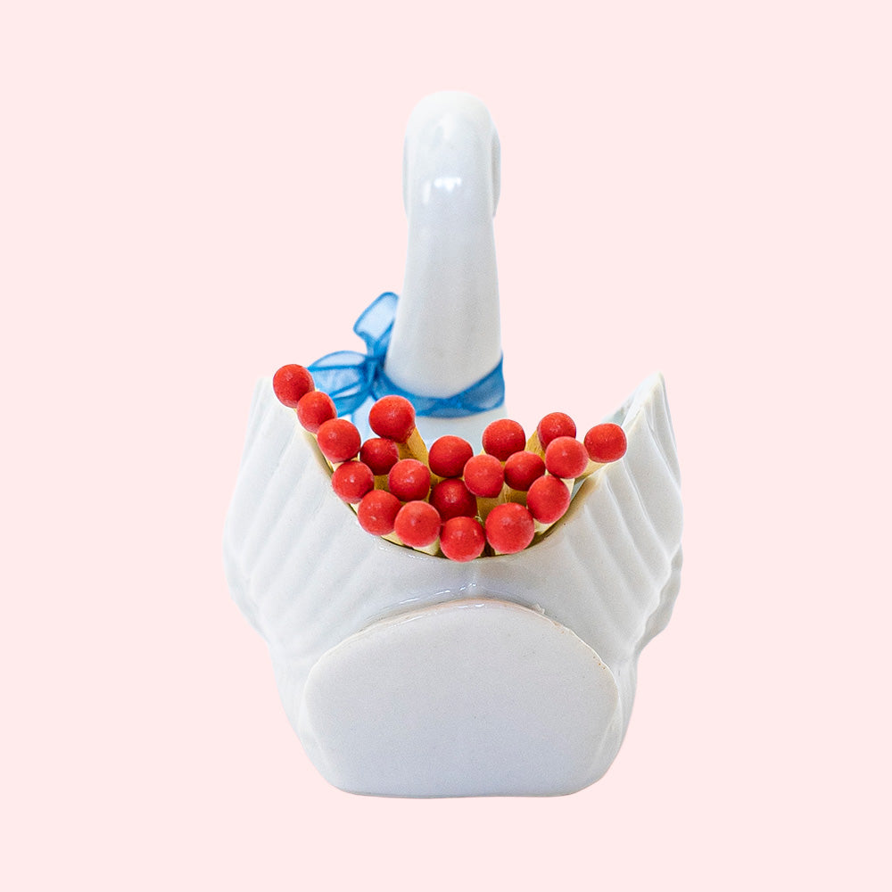 Second-hand ceramic, hollow swan figurine filled with 20 matches. It's wearing a blue organza ribbon.  Seen from the back.