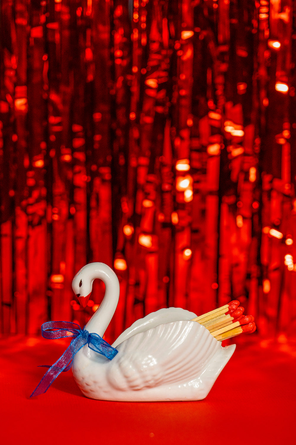 Second-hand ceramic, hollow swan figurine filled with 20 matches. It's wearing a blue organza ribbon. It sits on a bright red tinfoil background.