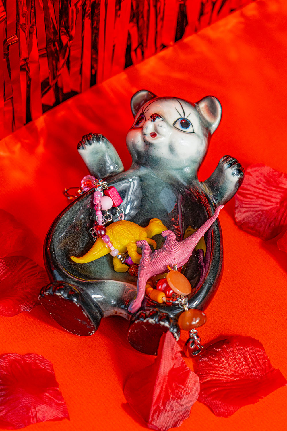 A Japan 1950's vintage trinket dish representing a black bear with a white face. In it are two handmade keychains made of beads and miniature dinosaur figurines. It lays on a bright red background with rose petals around it.