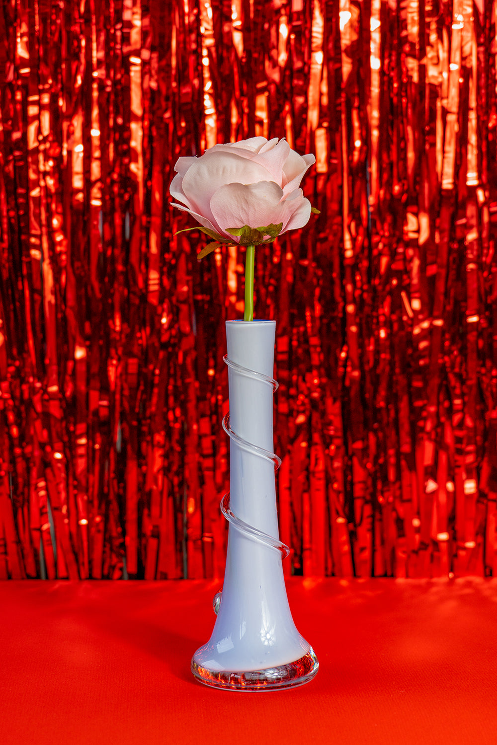 A vintage small blue opaline vase with a 3D swirl around it, holding a pink rose on a bright red tinfoil background.