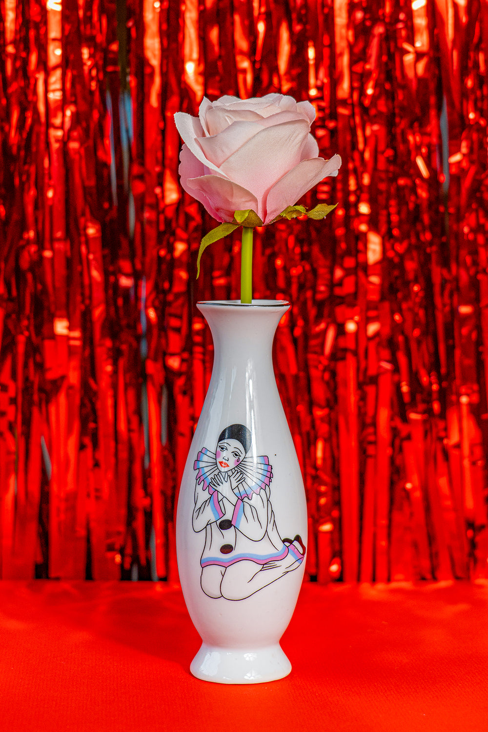White vase with Pierrot design and a black rim holding a pink rose against a red tinfoil background.