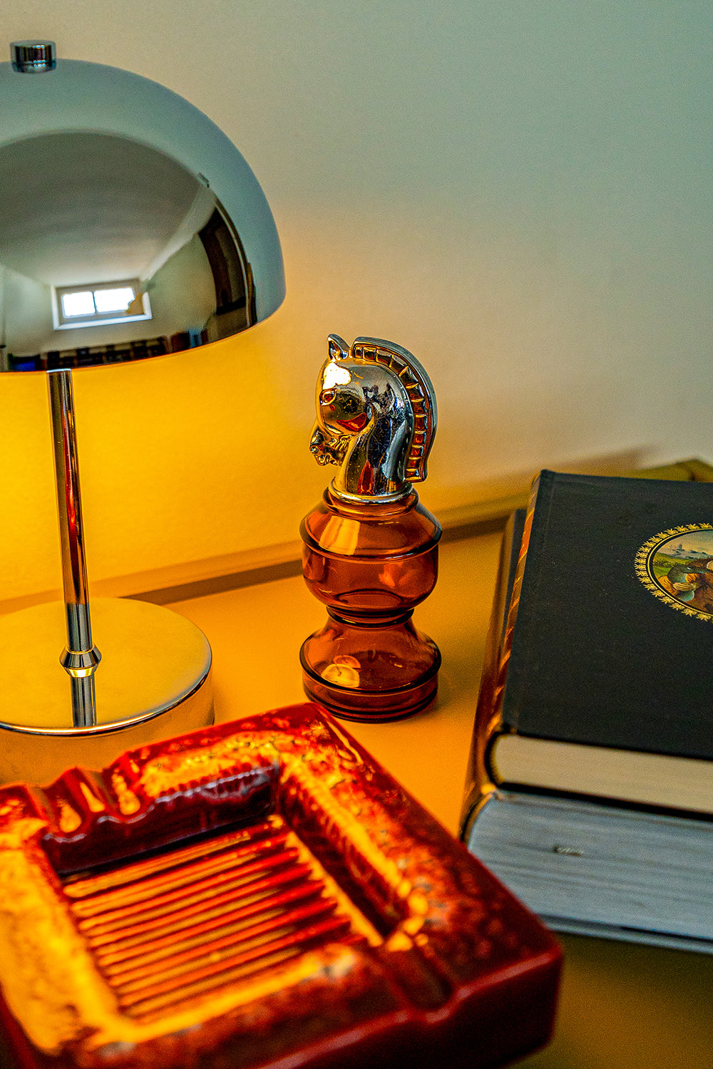 A decorative vintage bottle in amber glass with a silver horse head as the top, and a red square ashtray, sitting between a lamp and books